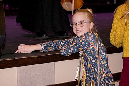 A young girl in a dress and glasses leans on the TCC Vantrease PACE stage, smiling as she enjoys the concert.