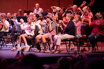 Signature Symphony at TCC musicians and audience members on stage, with American flags in the foreground.